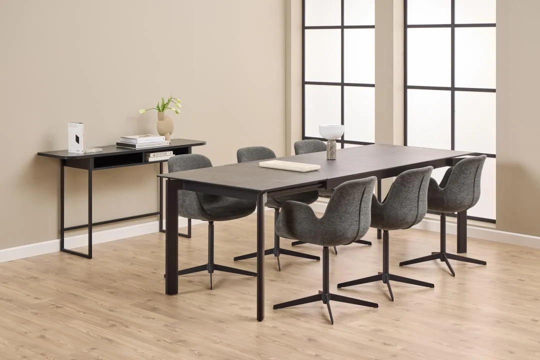 Een moderne eetkamer met de Eettafel Palermo 90 x 76 CM Rechthoek in robuust keramiek, omringd door zes grijze stoelen met kussens, een bijzettafel met boeken en een vaas, grote ramen voor natuurlijk licht, lichte houten vloeren en beige muren.