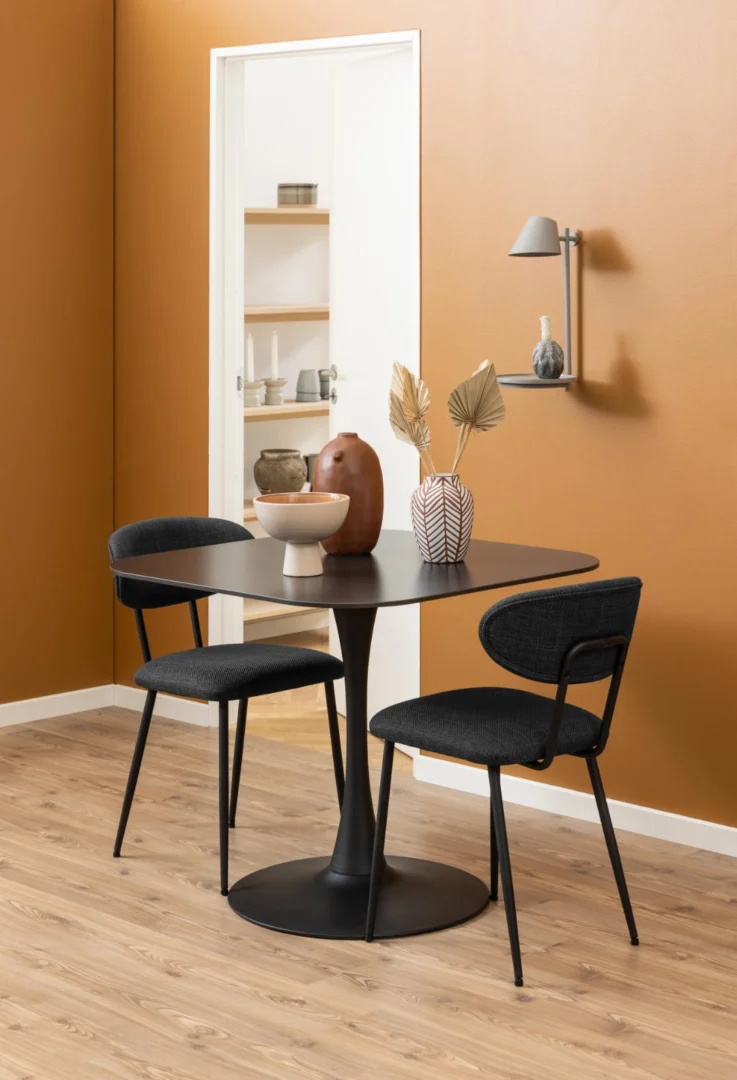 Een moderne eethoek met de Eettafel Nieuwe Sfeer 90 x 75 CM Rond (keramiek, staal), twee zwarte stoelen, decoratieve vazen met gedroogde planten, warme terracotta muren en aardewerk dat via een open deuropening op schappen staat uitgestald.