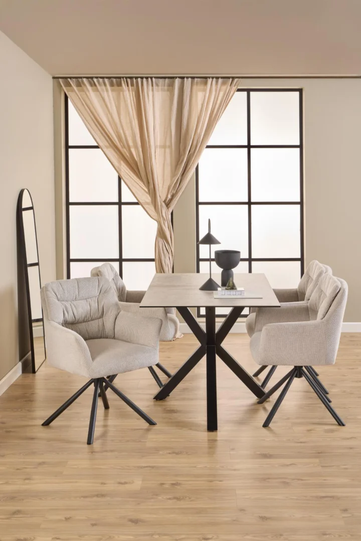 Een moderne eetruimte is voorzien van de Eettafel Nieuwe Sfeer 135 x 80 CM, een rechthoekige tafel van keramiek en staal, gekoppeld aan vier grijze stoelen met kussens, een kleine zwarte lamp, tafelbladdecoratie, transparante gordijnen bij grote ramen en een hoge spiegel op een beige muur.