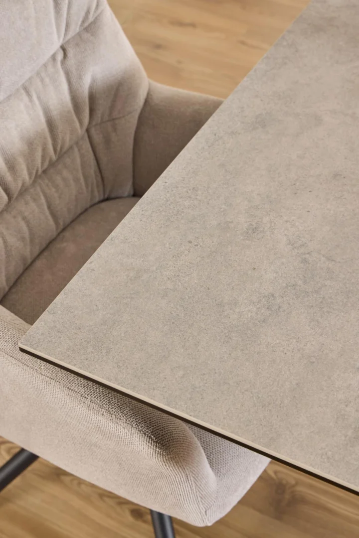 Een close-up van een beige gestoffeerde stoel naast de Eettafel Nieuwe Sfeer 135 x 80 CM, een rechthoekige tafel met een licht steenachtig keramiek oppervlak en een stalen frame, op een houten vloer.