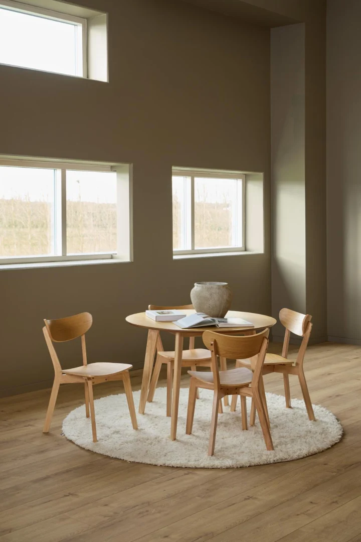 De Eettafel Nieuwe Sfeer (105 x 76 cm) in eikenhout staat met vier bijpassende stoelen op een wit vloerkleed in een zonovergoten kamer met grote ramen en lichte houten vloeren. Een decoratieve schaal en boeken sieren de tafel.