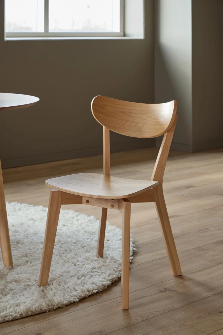 Een Eettafel Nieuwe Sfeer (105 x 76 cm) in eikenhout staat op een houten vloer naast een moderne houten stoel met een gebogen rugleuning, geplaatst op een rond wit kleed bij een groot raam.