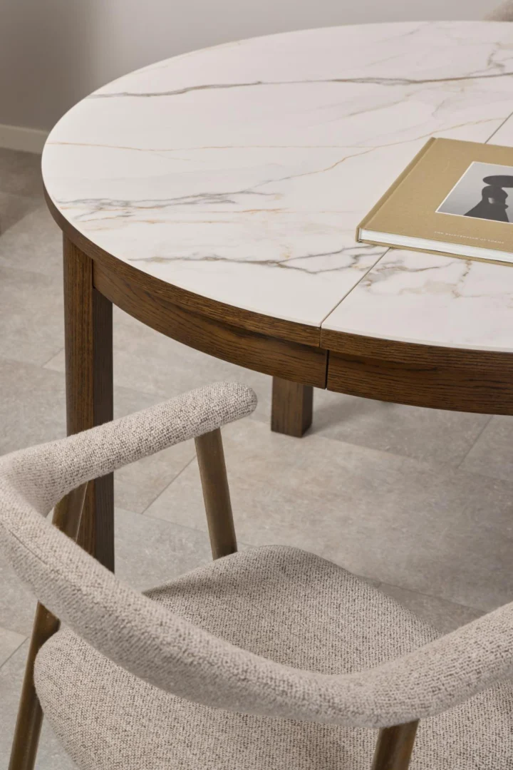 Een close-up van de Eettafel Nieuwe Sfeer 75 CM Rond Keramiek met een wit marmeren blad en donkerhouten rand, geplaatst naast een beige gestoffeerde stoel. Op de tafel ligt een dichtgeslagen boek met een zwart-wit foto.