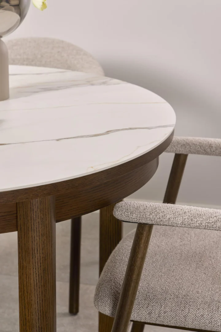 Een close-up van de Eettafel Nieuwe Sfeer 75 CM Rond Keramiek, met een rond keramisch blad met een houten rand, naast een lichtgrijs gestoffeerde stoel met houten poten en armleuningen.