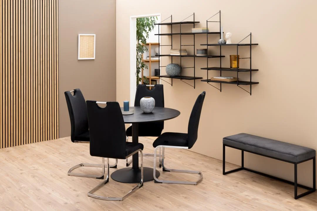 Een moderne eethoek met de Eettafel Nieuwe Sfeer 110 CM Rond MDF Mat met een zwart onderstel, vier zwarte stoelen, een zwarte wandplank met decor, een grijze bank, lichthouten vloeren en een deuropening die weelderig groen onthult.