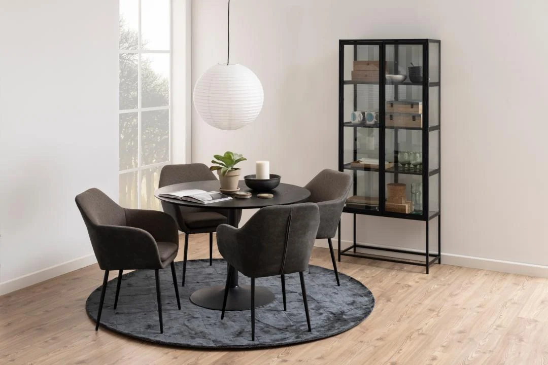 Een moderne eethoek met de Eettafel Nieuwe Sfeer 110 CM Rond MDF Mat met zwart onderstel, vier grijze stoelen, een rond vloerkleed, een zwarte vitrinekast met decor, een ronde hanglamp en een houten vloer bij een raam.