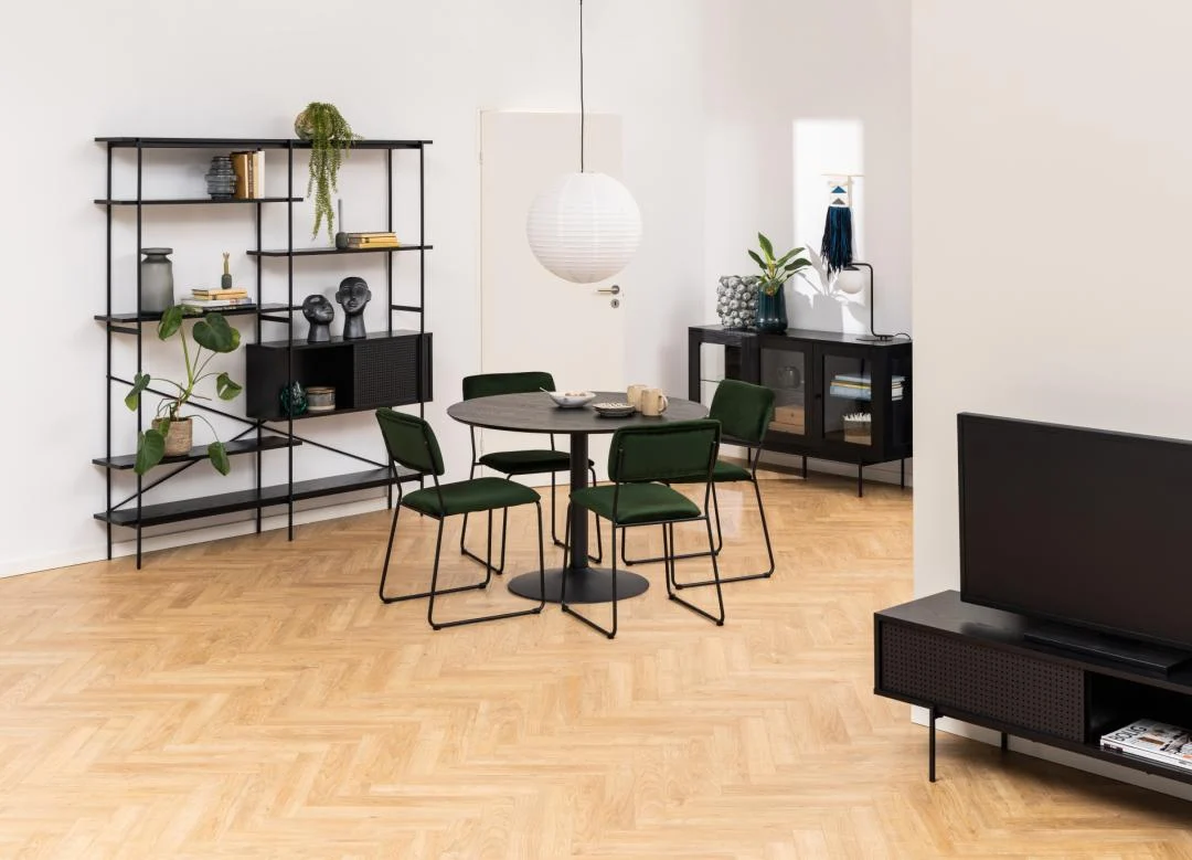 Een moderne woon- en eetkamer met houten visgraatvloer, de Eettafel Nieuwe Sfeer 110 CM Rond MDF Mat met een zwarte basis, groene stoelen, zwarte planken met decor, een zwart TV-meubel en minimalistische accessoires tegen witte muren.