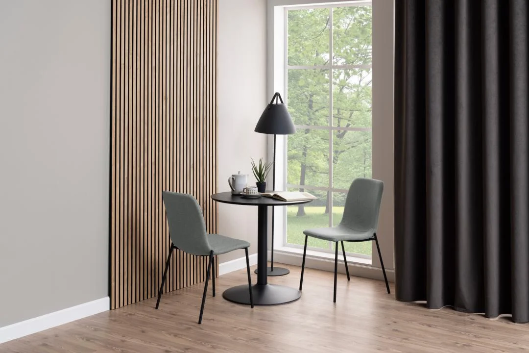 De Eettafel Nieuwe Sfeer 80 CM Rond MDF Mat met zwart onderstel staat met twee grijze stoelen bij een groot raam met uitzicht op groene bomen. Boven de tafel hangt een zwarte lamp; de kamer heeft een houten vloer, een lattenwand en donkere gordijnen.