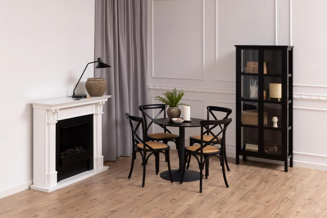 Een moderne eethoek met de Eettafel Nieuwe Sfeer 80 CM Rond MDF Mat met zwart onderstel, vier zwarte stoelen, een plantenpot, witte open haard, zwarte vitrinekast, houten vloer en lichtgrijze gordijnen op witte muren.