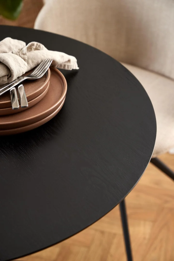 Een close-up van de Eettafel Nieuwe Sfeer 80 CM Rond MDF Mat met zwart onderstel, set met gestapelde bruine borden, twee vorken en een beige servet naast een licht gestoffeerde stoel op een houten vloer.