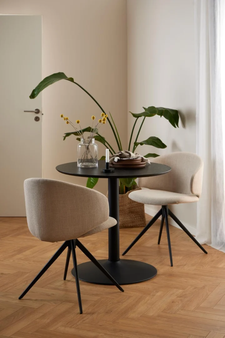 Een lichte eethoek met de Eettafel Nieuwe Sfeer 80 CM Rond MDF Mat met zwart onderstel, twee beige gestoffeerde stoelen, een glazen vaas met gele bloemen, boeken en een potplant op een houten vloer met neutrale muren.