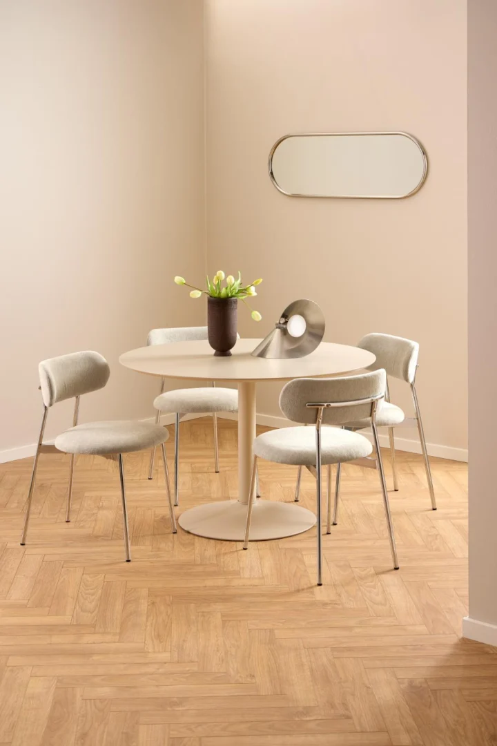 Een moderne eethoek met de Eettafel Nieuwe Sfeer 110 CM Rond Mdf, vier witte stoelen met metalen poten, een vaas met witte tulpen, een metalen decorstuk, een visgraat vloer van licht hout, beige muren en een ovale spiegel.