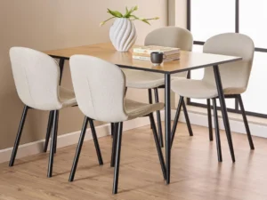 Een moderne eethoek met de Eettafel Wilma 80 x 75 CM ronde MDF tafel met een mat zwart stalen onderstel, vier beige stoelen met kussens en zwarte poten, een witte vaas met bloemen, drie gestapelde boeken en een zwarte mok op een licht houten vloer bij grote ramen.