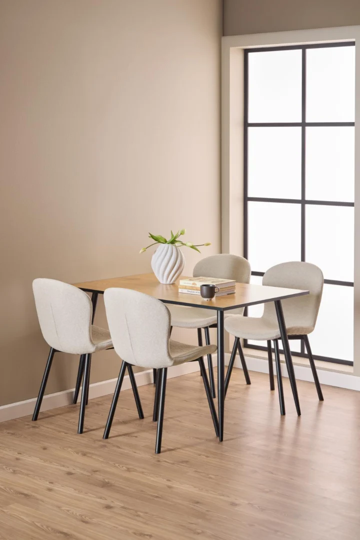 Een moderne eethoek is voorzien van de Eettafel Wilma 80 x 75 CM ronde MDF tafel met een matzwart stalen onderstel, vier beige gestoffeerde stoelen, een vaas met bloemen, gestapelde boeken, een mok, een lichthouten vloer en een groot raam met zwarte omlijsting.