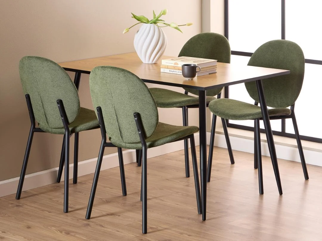 Een moderne eethoek met de Eettafel Wilma 80 x 75 CM ronde MDF tafel met mat zwart onderstel, vier groen gestoffeerde stoelen met zwarte poten, een witte vaas met groen, boeken en een mok op een licht houten vloer bij een groot raam.