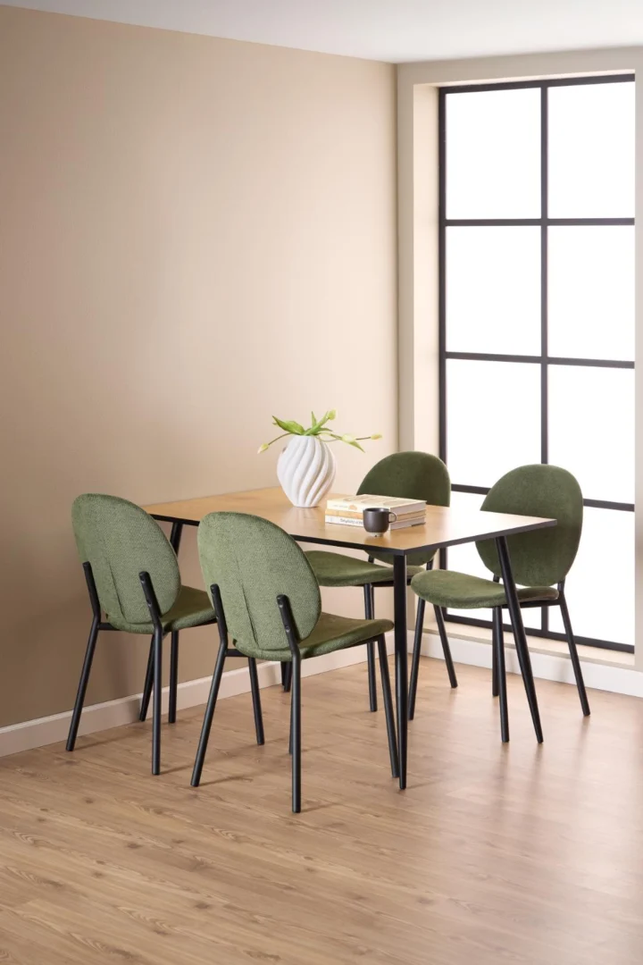 Een stijlvolle eetruimte met de Eettafel Wilma 80 x 75 CM rond MDF mat met zwart onderstel, gecombineerd met vier groen gestoffeerde stoelen, een witte vaas met bloemen, een zwarte mok en twee boeken op een licht houten vloer bij een groot raam.