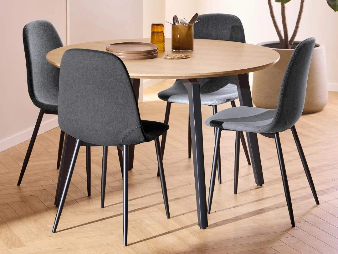 De Eettafel 1 (120 cm rond, MDF, mat zwart onderstel) wordt getoond met vier grijs gestoffeerde stoelen, gedekt met borden, glazen en keukengerei op een lichthouten vloer naast een grote potplant.