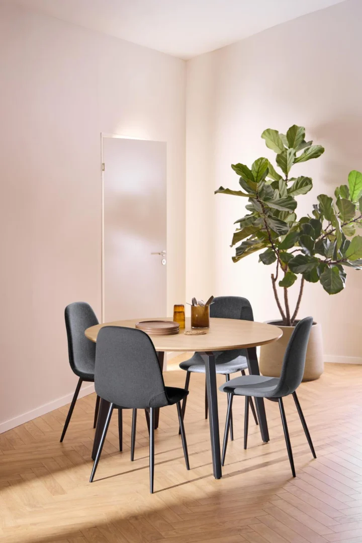 Een ronde Eettafel 1 120 CM met MDF blad en zwart stalen onderstel is gekoppeld aan vier grijze stoelen op een visgraat houten vloer. Bruine glazen voorwerpen staan op de tafel, met een grote, bebladerde potplant bij een beige muur en gesloten deur.
