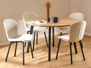 De Eettafel 1 120 CM Rond MDF met een matzwart stalen onderstel wordt omringd door vier beige gestoffeerde stoelen. Op de tafel staan een theepot, kopje, open boek en geweven lamp, allemaal op een lichte houten vloer.