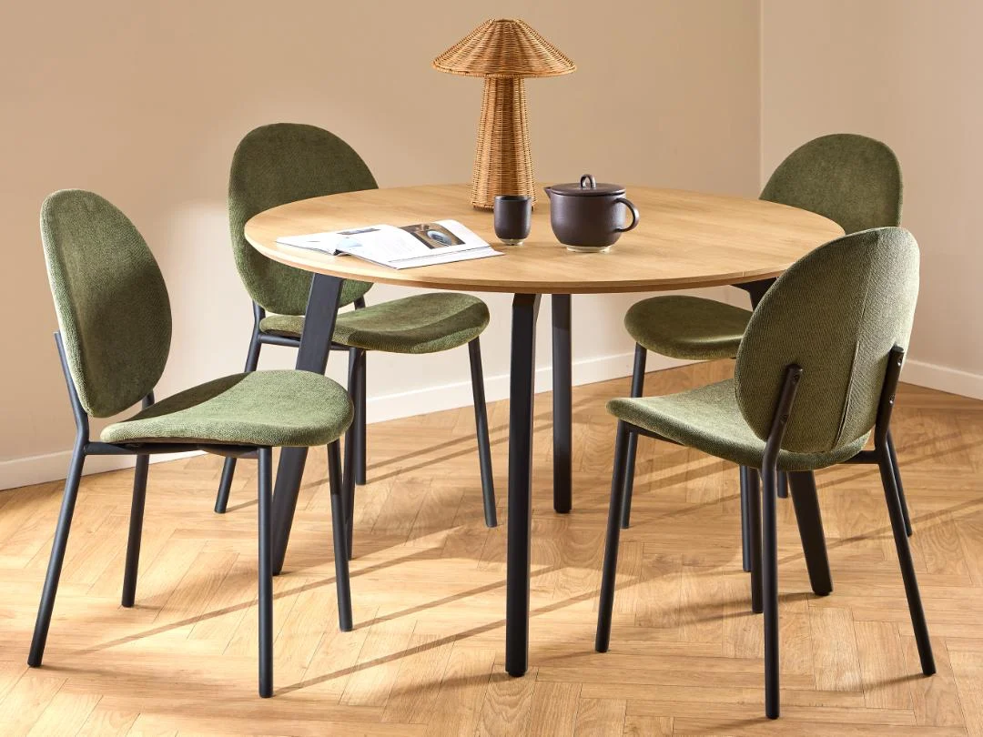 De Eettafel 1 120 CM Rond Eikenhout met matzwarte stalen poten is gecombineerd met vier groen gestoffeerde stoelen met zwart onderstel, gestyled in een lichte moderne kamer met lichte vloer- en tafeldecoratie.