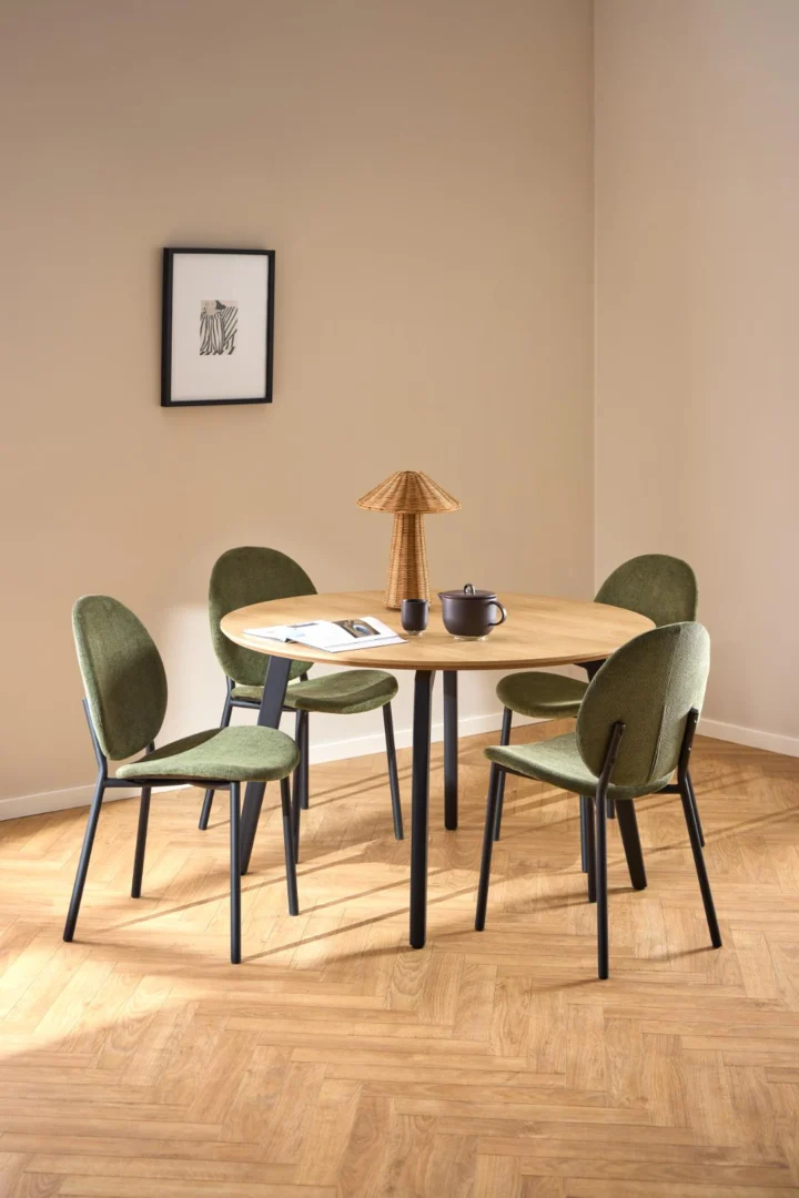 De Eettafel 1 120 CM Rond Eikenhout Staal met een mat zwart onderstel staat in een minimalistische kamer met een houten vloer, beige muren, een ingelijst kunstwerk en wordt omringd door vier groene stoelen, met daarop een lamp, theepot, kopje en open boek.