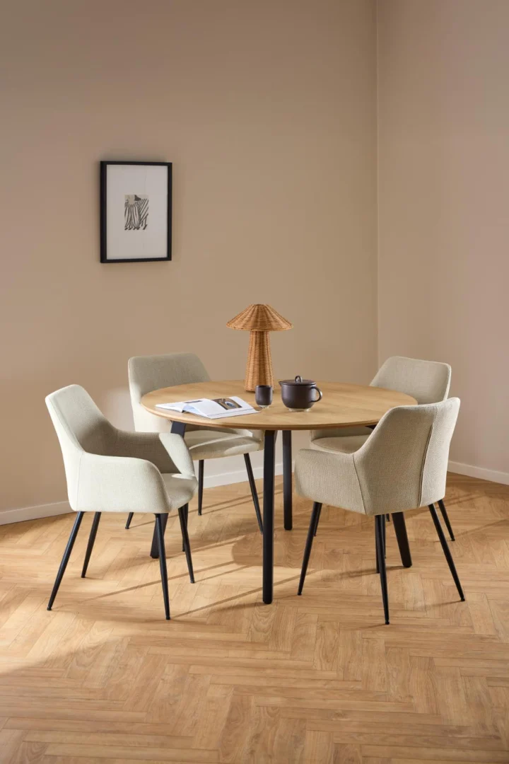 Een ronde eiken eettafel van 120 cm (Eettafel 1) met een matzwart stalen onderstel, gekoppeld aan vier beige stoelen en afgewerkt met een lamp met geweven kap en een theepot. De kamer heeft lichtbruine muren, een houten vloer en ingelijste muurkunst.
