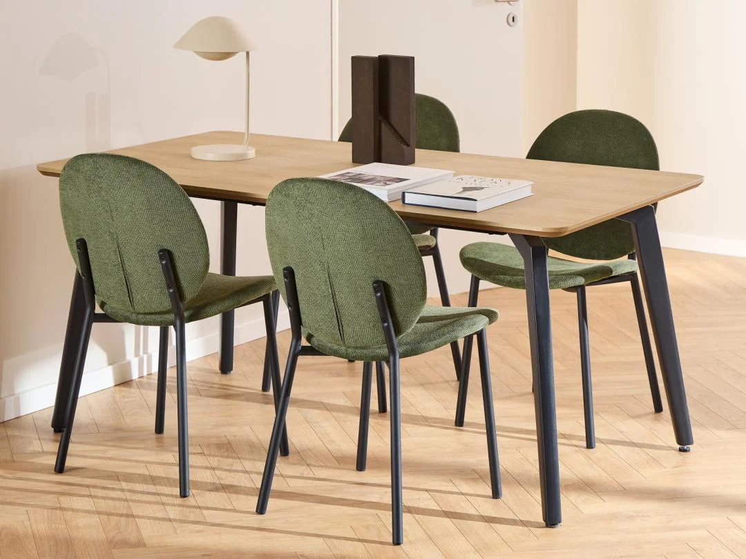 De Eettafel Nieuwe Sfeer 90 x 75 CM Rond, gemaakt van eikenhout, staat op een lichthouten visgraatvloer met vier groen gestoffeerde stoelen, een beige lamp, decoratieve boekensteunen en twee boeken erop.