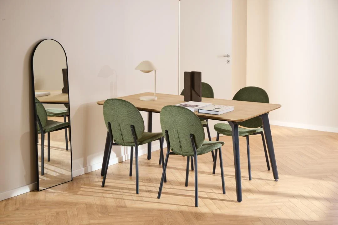 In een moderne eetkamer staan de Eettafel Nieuwe Sfeer 90 x 75 CM Rond in eikenhout, vier groen gestoffeerde stoelen, een tafellamp, boeken, decor en een hoge gewelfde spiegel tegen een beige muur op een visgraat houten vloer.