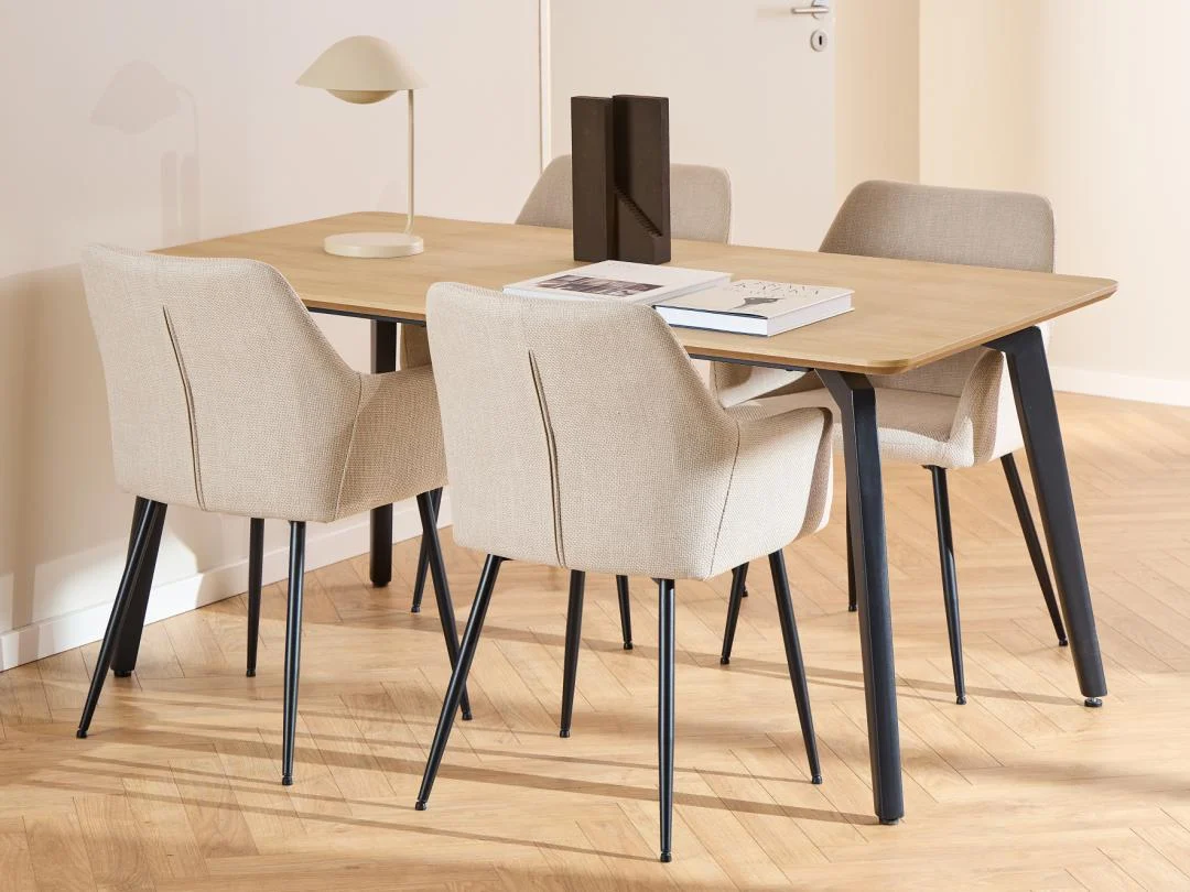 Een moderne eetkamer met de Eettafel Nieuwe Sfeer 90 x 75 CM ronde MDF en stalen tafel, vier beige gestoffeerde stoelen met zwarte poten, boeken en een lamp op de tafel, op een lichthouten visgraatvloer.