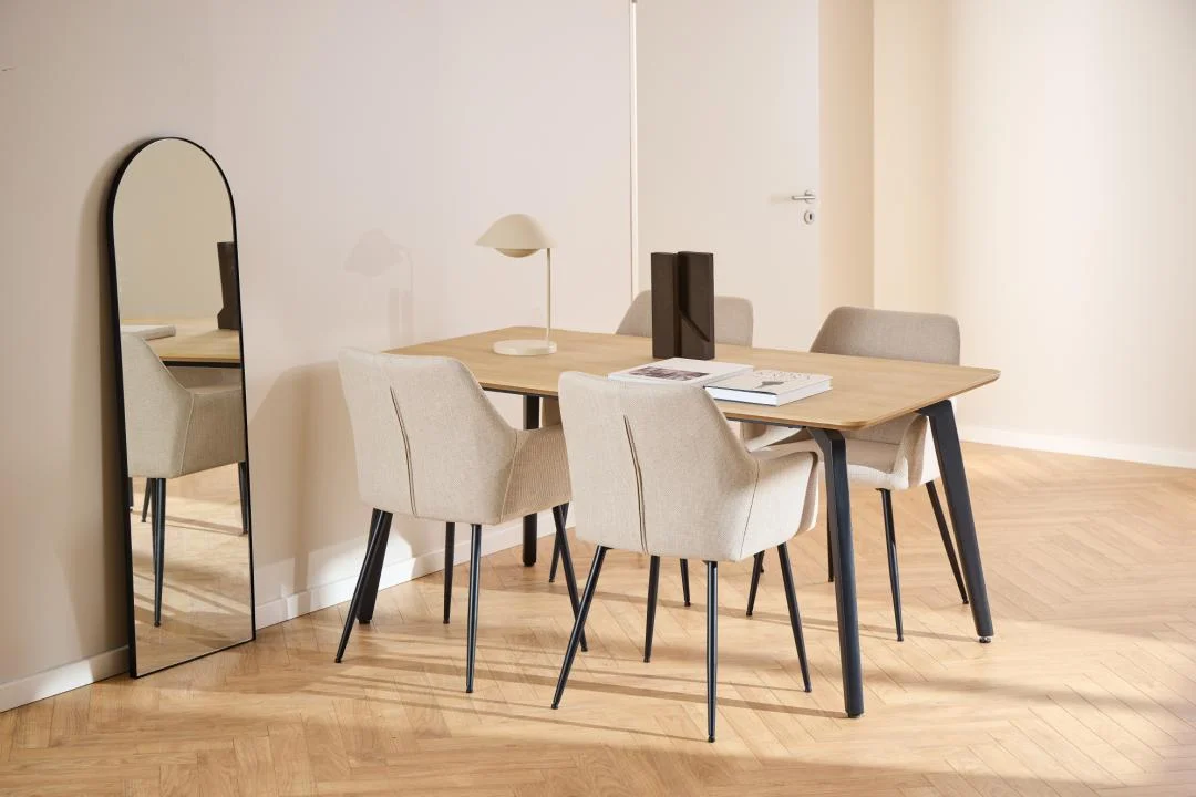 Een moderne eetkamer met de Eettafel Nieuwe Sfeer 90 x 75 CM Rond MDF Staal tafel, vier beige gestoffeerde stoelen, een kleine lamp, boeken en een decoratief stuk. Een hoge gewelfde spiegel leunt tegen de lichte muur; lichte visgraat houten vloeren maken de look compleet.