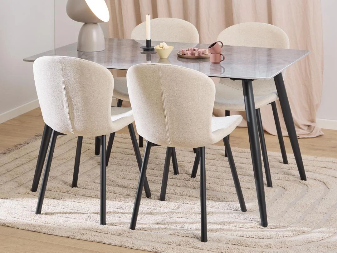 Een moderne eethoek met de Eettafel Nieuwe Sfeer, een ronde uittrekbare tafel van keramiek van 80 x 75 cm, omringd door vier gestoffeerde crèmekleurige stoelen met zwarte poten, een vloerkleed en minimalistische decoraties zoals een lamp, een kaars, een schaal en een roze schaal.