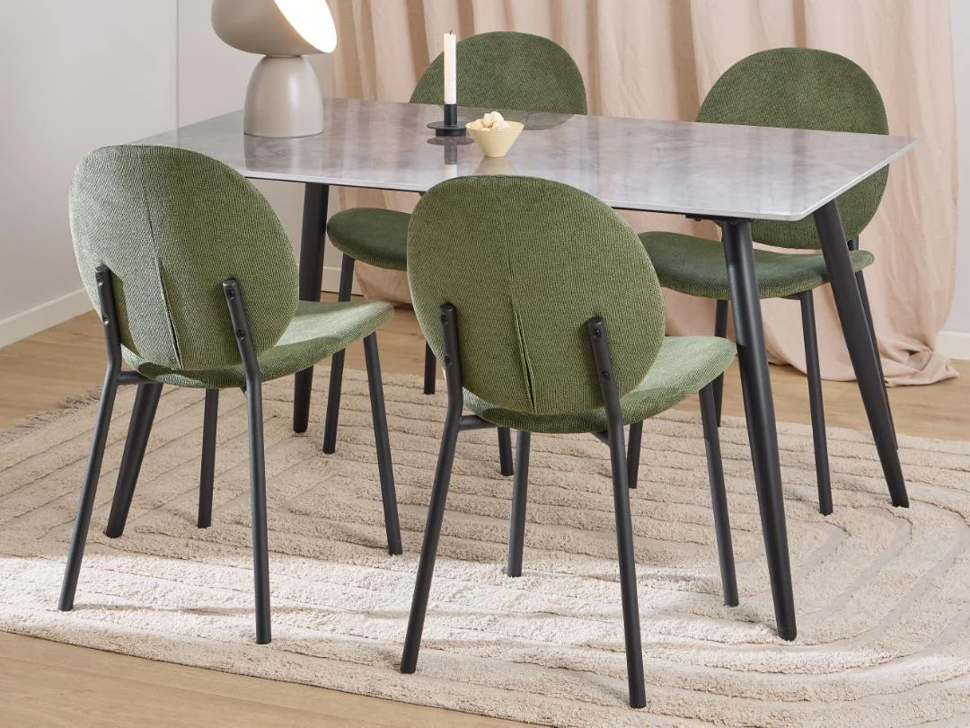 Een moderne eethoek met de ronde Eettafel Wicklow 80 x 75 CM in zwart keramiek, vier groen gestoffeerde stoelen met zwarte poten en een beige vloerkleed op een houten vloer. De tafel is gestyled met een lamp, kaars en decor; beige gordijnen maken de look compleet.