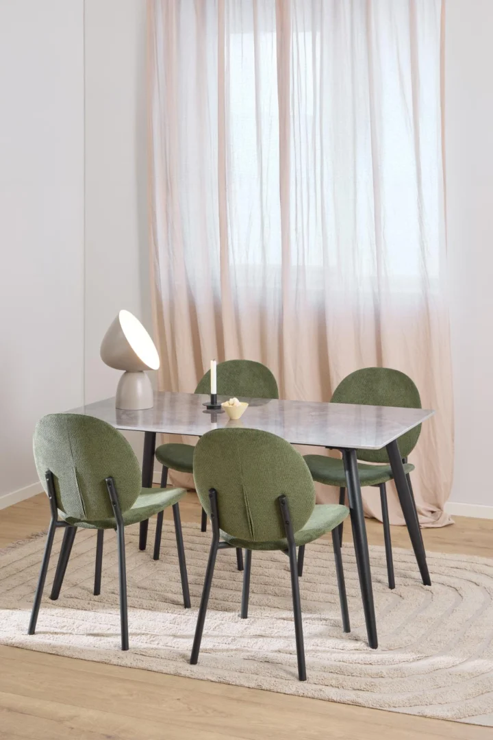 Een moderne eethoek met de Eettafel Wicklow 80 x 75 CM Rond Keramiek in zwart, vier groen gestoffeerde stoelen, een beige vloerkleed, een sculpturale lamp en kaarsen. Gordijnen bedekken het raam en laten zacht natuurlijk licht binnen.