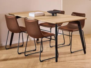 De Eettafel Nieuwe Sfeer 90 x 75 CM Rond, gemaakt van eikenhout en staal, wordt omringd door vier bruine leren stoelen met metalen frames. Op de eettafel staan twee boeken en een zwart decoratief object.