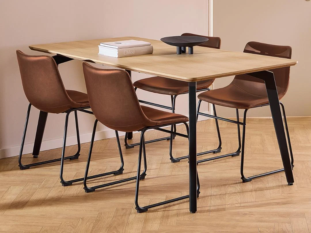 De Eettafel Nieuwe Sfeer 90 x 75 CM Rond, gemaakt van eikenhout en staal, wordt omringd door vier bruine leren stoelen met metalen frames. Op de eettafel staan twee boeken en een zwart decoratief object.