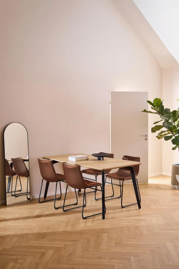 Een moderne eethoek met de Eettafel Nieuwe Sfeer 90 x 75 CM Rond eikenhout staal tafel met vier bruine stoelen, een hoge vloerspiegel, een potplant en een witte deur op een lichthouten visgraatvloer in zacht natuurlijk licht.