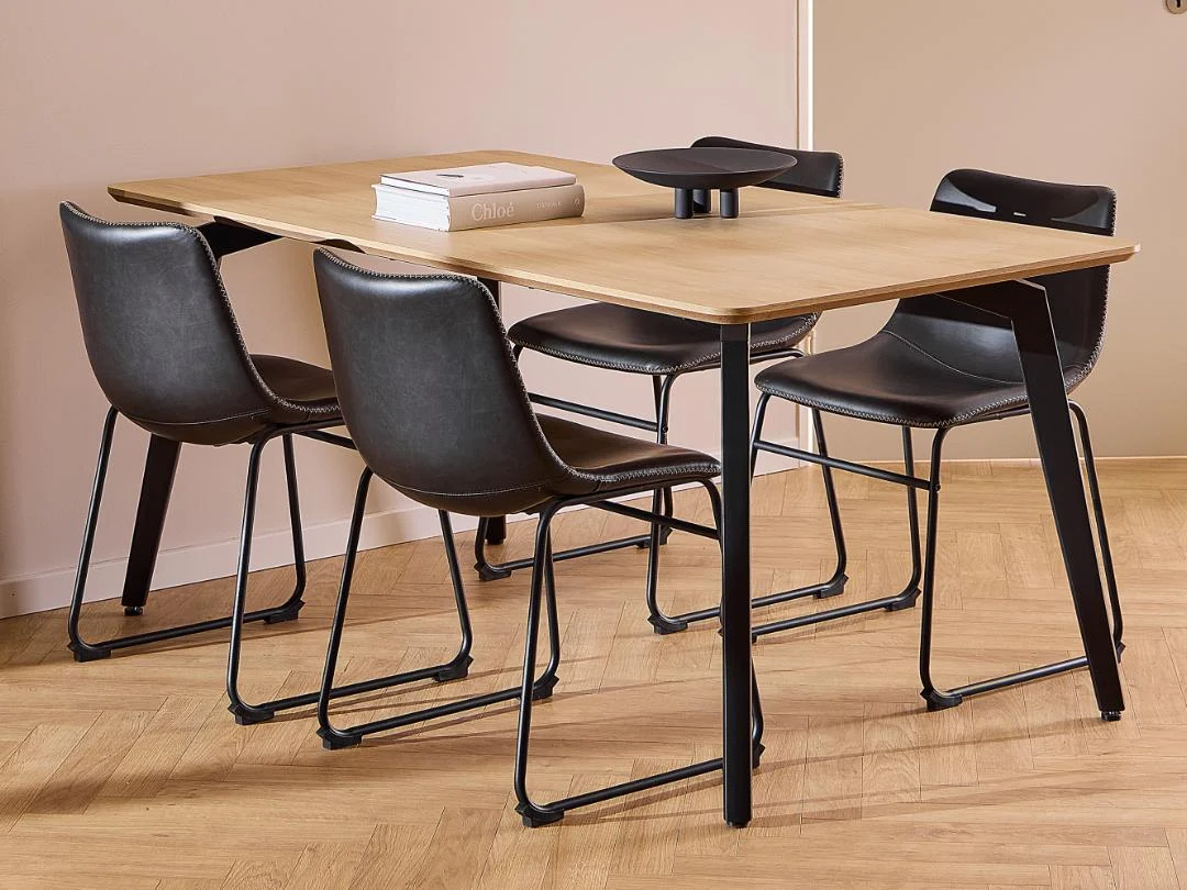 De Eettafel Nieuwe Sfeer (90 x 75 CM, rond) heeft een licht eikenhout oppervlak en zwarte stalen poten, gecombineerd met vier zwartleren stoelen met metalen frames. Op de tafel liggen twee boeken en een zwart decoratief object, op houten vloer.