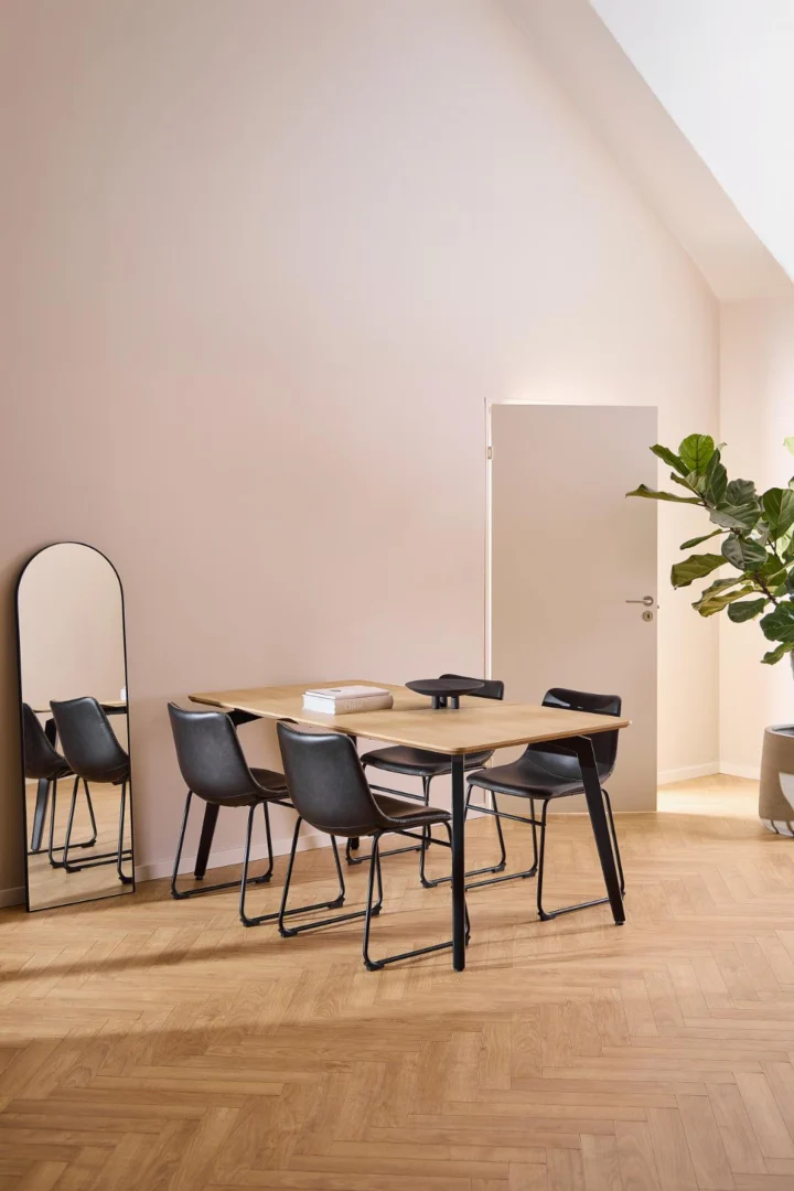 Een minimalistische eethoek met de Eettafel Nieuwe Sfeer 90 x 75 CM Rond in eikenhout staal, zes zwarte stoelen, een grote plant en een hoge spiegel, tegen bleke muren en een visgraat houten vloer met natuurlijk licht.