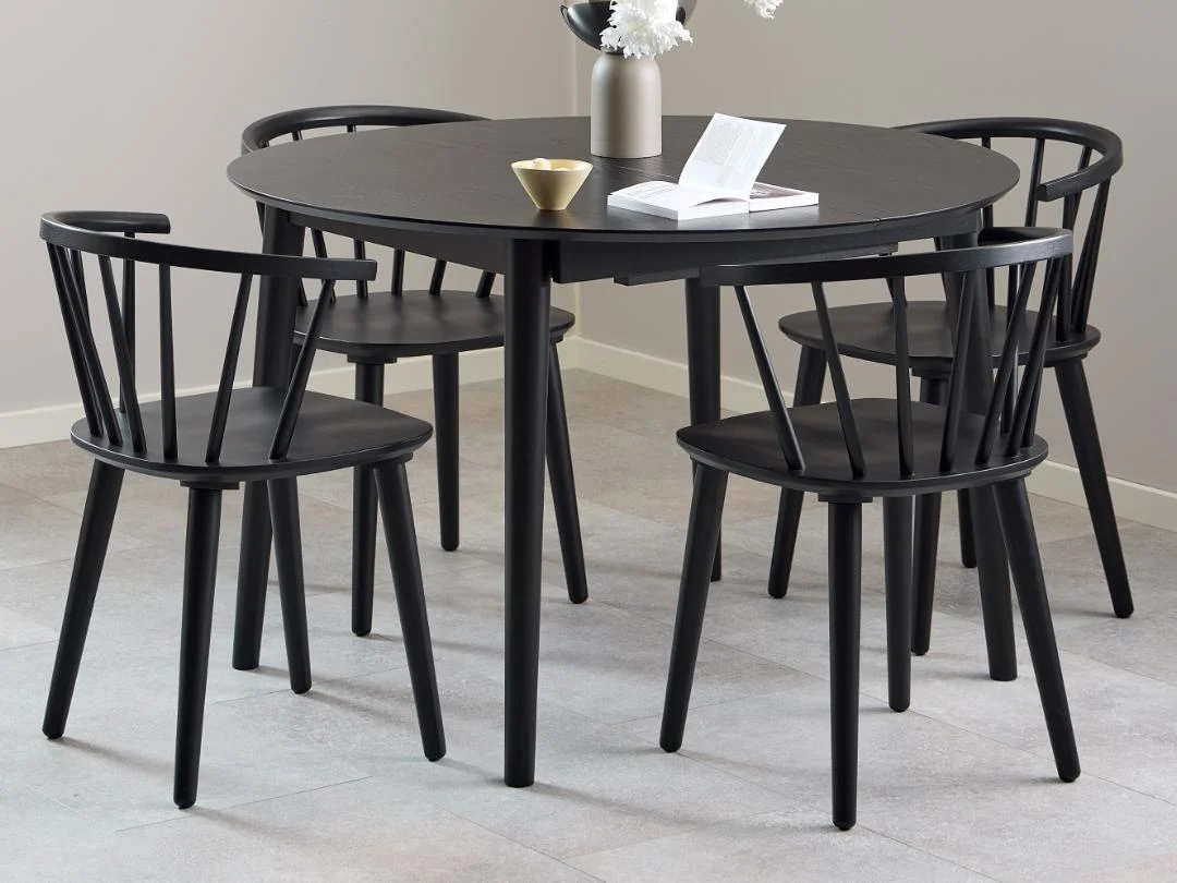 De Eettafel Nieuwe Sfeer 115 CM Rond MDF Mat met een zwart onderstel is vormgegeven met vier bijpassende zwarte stoelen, een vaas met witte bloemen, een open boek en een klein gouden kopje bovenop het oppervlak, allemaal geplaatst op lichte tegels in een minimalistische kamer.