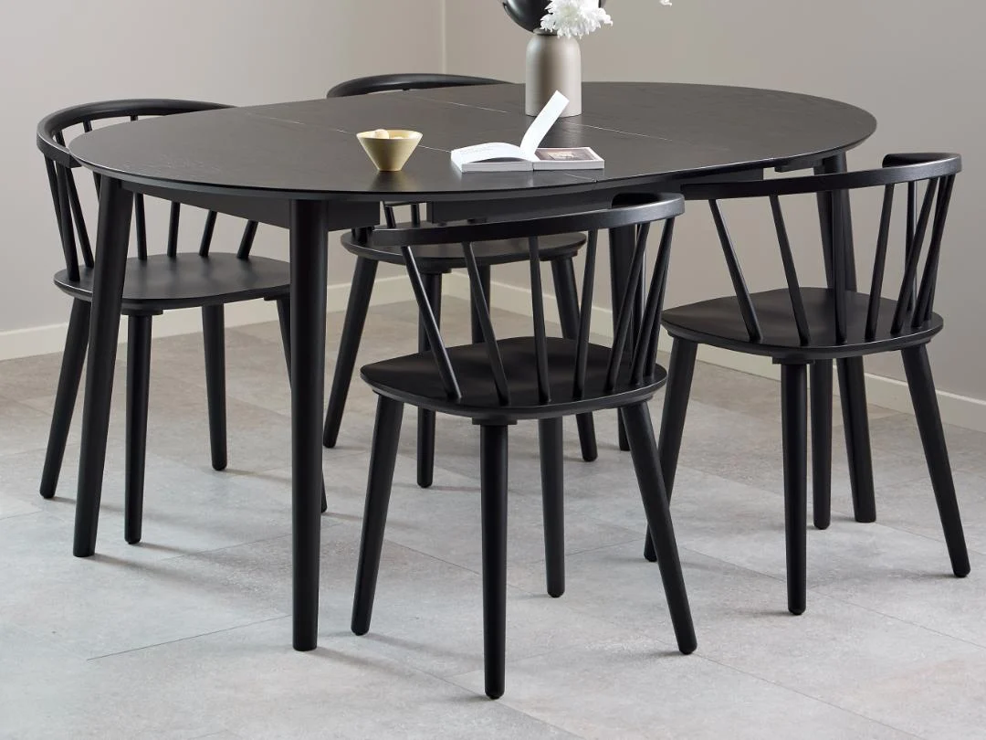 Een moderne eetset met de Eettafel Nieuwe Sfeer 115 CM Rond MDF Mat met een zwart onderstel, gekoppeld aan vier bijpassende zwarte stoelen. De tafel is gestyled met een bloemenvaas, een open boek en een gouden schaal. De kamer heeft een lichte tegelvloer en effen muren.