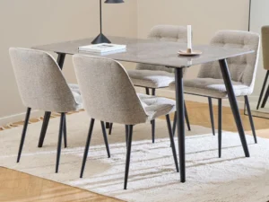 Een moderne eethoek met de Eettafel Wicklow 80 x 75 CM Rechthoek Mat met zwart onderstel, vier gestoffeerde beige stoelen met zwarte poten, een boek, kaars en lamp op tafel, boven een licht vloerkleed op een houten vloer. Een spiegel weerspiegelt het tafereel.