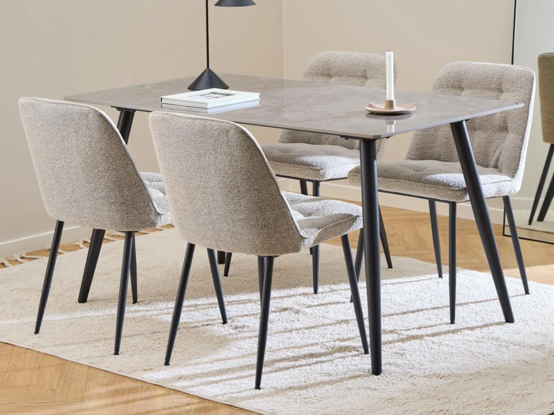 Een moderne eethoek met de Eettafel Wicklow 80 x 75 CM Rechthoek Mat met zwart onderstel, vier gestoffeerde beige stoelen met zwarte poten, een boek, kaars en lamp op tafel, boven een licht vloerkleed op een houten vloer. Een spiegel weerspiegelt het tafereel.