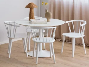 De Eettafel Ibiza 110 CM Rond Mdf, een ronde witte eettafel, staat met vier bijpassende stoelen op een lichte houten vloer. Het is gedecoreerd met een lamp, gele bloemen in een vaas en een boek, terwijl beige gordijnen op de achtergrond hangen.