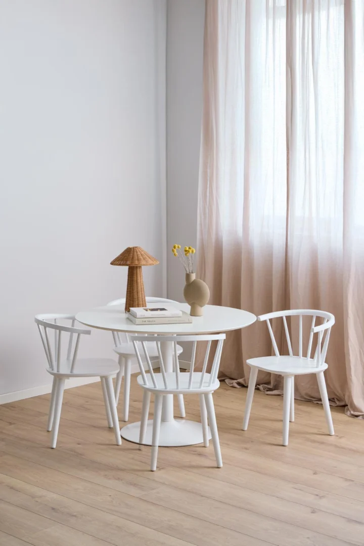 Een minimale eethoek is voorzien van de Eettafel Ibiza 110 CM Rond Mdf tafel, drie witte stoelen, een beige vaas met gele bloemen, boeken en een geweven lamp. Gordijnen laten zonlicht de zacht verlichte kamer met houten vloeren vullen.