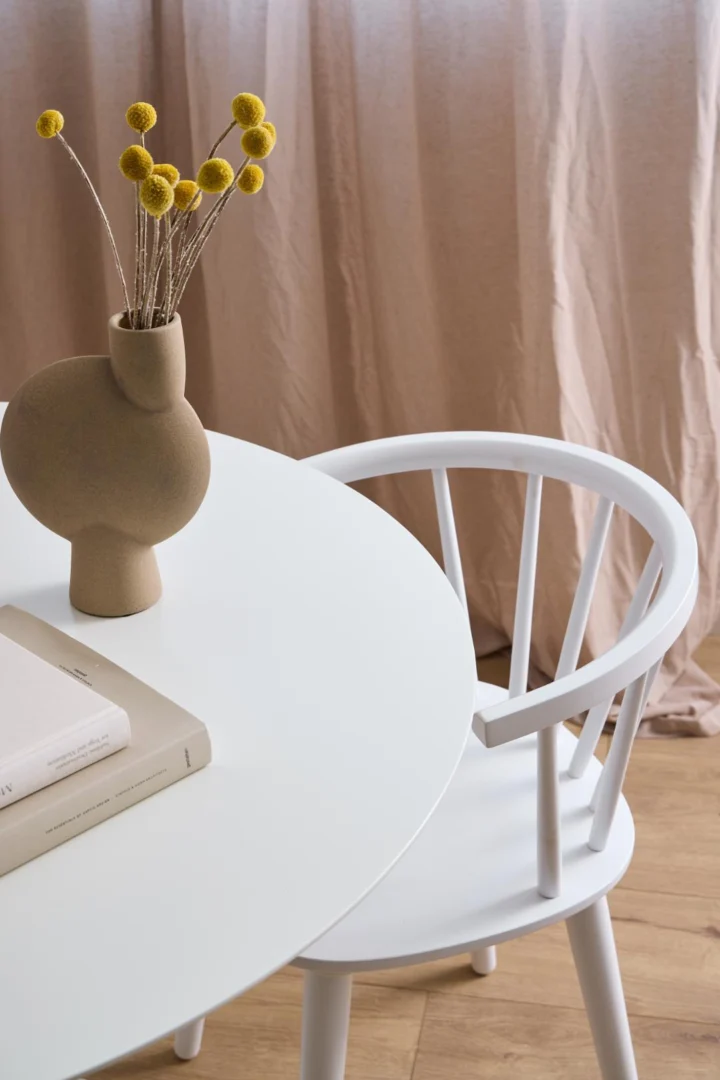 Een minimalistisch tafereel met de Eettafel Ibiza 110 CM Rond MDF, een bijpassende witte stoel, een beige sculpturale vaas met gele bollen en twee gesloten boeken, tegen beige gordijnen en een houten vloer.