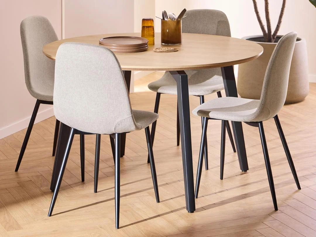 De Eettafel 1 120 CM Rond Eikenhout Staal met mat zwart onderstel staat met vier beige gestoffeerde stoelen op een lichte visgraat vloer. Borden, glazen en keukengerei staan netjes op tafel, met een grote plant op de achtergrond.