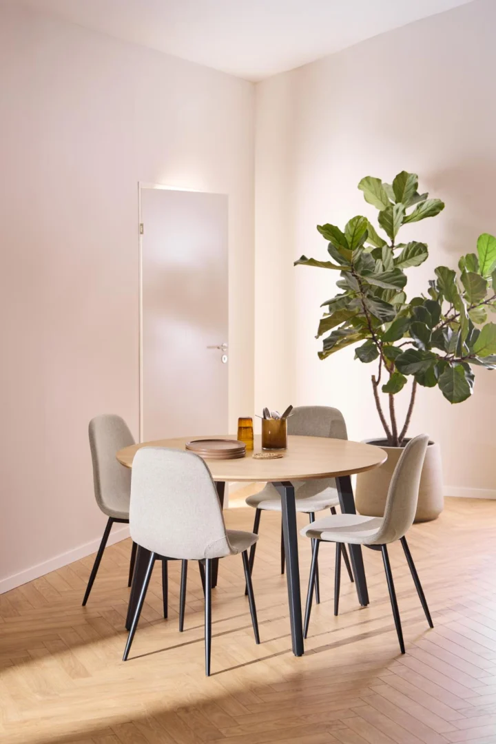 Een moderne eethoek met de Eettafel 1 120 CM Rond Eikenhout Staal met een mat zwart onderstel, vier lichtgrijze stoelen met zwarte poten, twee amberkleurige glazen kopjes, een grote potplant in de hoek en een lichthouten visgraatvloer.