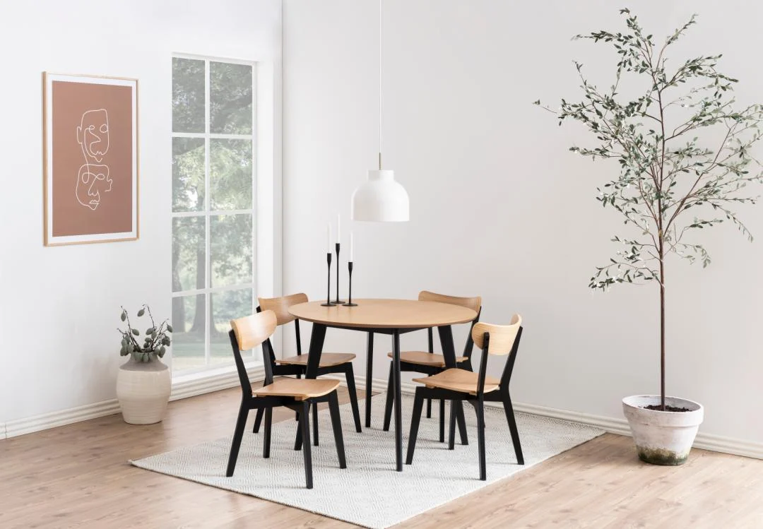 Een minimalistische eetkamer met de Eettafel Nieuwe Sfeer 105 CM Rond in eikenhout, vier bijpassende stoelen, een witte hanglamp, potplanten, abstracte muurkunst en een groot raam dat natuurlijk licht binnenlaat.