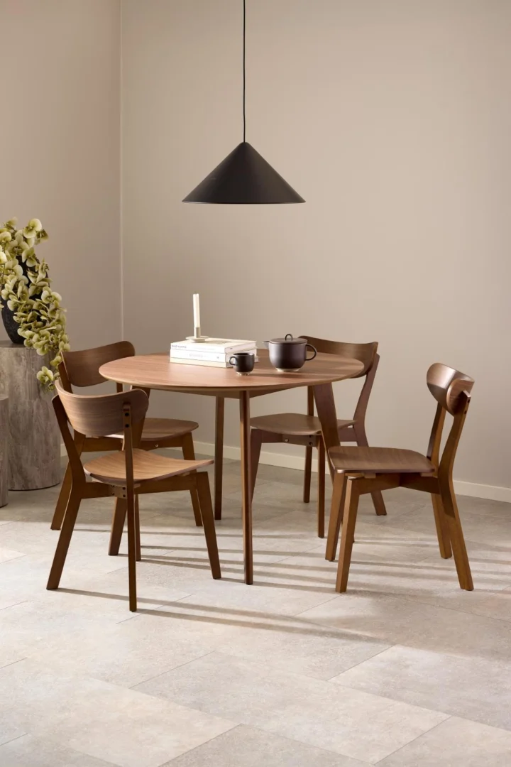 In een moderne eethoek staat de Eettafel Roxby 105 CM Rond Notenhout, omringd door vier houten stoelen. Daarboven hangt een zwarte hanglamp. Op de tafel staan een theepot, kopjes, een kaars en een boek; in de hoek staat een vaas met groene bladeren.