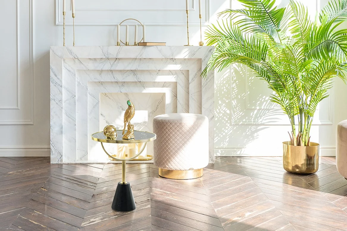 Een lichte, elegante woonkamerhoek met een witte marmeren open haard, een Bijzettafel Zwart Marmer Compact, een gewatteerde poef en een grote palmplant in pot op een vloer van donker visgraathout.